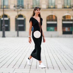 Amanda Derhy wears earrings, a black low-neck v-neck dress, a white circular round straw summer bag, a watch, white Adidas sneakers shoes, on August 09, 2020 in Paris, France.