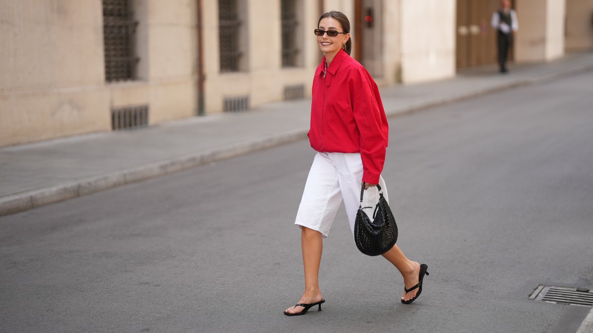 MUNICH, GERMANY - JUNE 8: Lina Uhrig is seen wearing a red cotton cropped jacket with a shirt collar, front zip closure, long sleeves, relaxed oversized silhouette and minimal design from Mango x Hailey Bieber; white cotton Bermuda shorts with a straight silhouette, knee length, clean waistband and minimal design from Stradivarius; black leather kitten heel sandals with thin toe straps, open toe construction and low slim heels from Stradivarius; a natural pedicure visible through the sandals; a black woven leather shoulder bag with a compact curved silhouette, short top handles, textured woven surface and metal hardware details from Adax; gold tone hoop earrings; rectangular black acetate sunglasses with narrow frames and dark tinted lenses from Chimi; her brown hair is worn in a sleek low ponytail with a center part; with natural make up on June 8, 2026 in Munich, Germany. (Photo by Moritz Scholz/Getty Images)