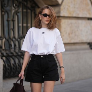 MUNICH, GERMANY - JUNE 8: Pia Riegel is seen wearing an oversized white t-shirt by Adidas; black high-waisted denim shorts by Agolde; a dark brown suede wrist bag with a soft slouchy shape and short rounded handles by COS; black oval sunglasses by Ray-Ban; silver headphones by Apple; a gold-tone wristwatch with a light dial and black leather strap; gold hoop earrings; shoulder-length softly tousled hair; with natural make up on June 8, 2026 in Munich, Germany. (Photo by Moritz Scholz/Getty Images)