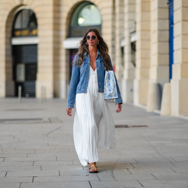 Amanda Derhy wears earrings, sunglasses, a blue denim jacket, a white summer maxi pleated low-neck slit off-shoulder dress, sandals, during a street style fashion photo session, on August 20, 2022 in Paris, France.