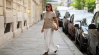 Diane Batoukina wears long straight brown hair, black rectangular sunglasses by Montblanc, a beige chunky knit short-sleeve top by Massimo Dutti, a white maxi skirt by Massimo Dutti, a beige clutch bag by Flattered, dark brown sandals leather shoes by Soeur, during a street style fashion photo session, on June 16, 2026 in Paris, France.