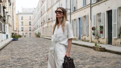 PARIS, FRANCE - MAY 07: Natalia Verza wears long straight light brown hair with blonde highlights and a center part, oversized black sunglasses with brown tinted lenses and thick frames, natural makeup with nude lipstick, a delicate silver-tone necklace, a white Munthe short-sleeve shirt with thin vertical gray stripes, an open collar, front button fastening, gold-tone buttons, a loose fit and a slightly uneven hem, white Munthe bermuda shorts with matching thin vertical gray stripes, a high waist, and knee-length cut, a dark brown leather Munthe bag with two short handles, a soft slouchy shape and tonal front logo detail, and brown Potamy slingback pumps with pointed toes, slim ankle straps, small kitten heels and a suede-effect finish, during a street style fashion photo session, on May 07, 2026 in Paris, France. (Photo by Edward Berthelot/Getty Images)