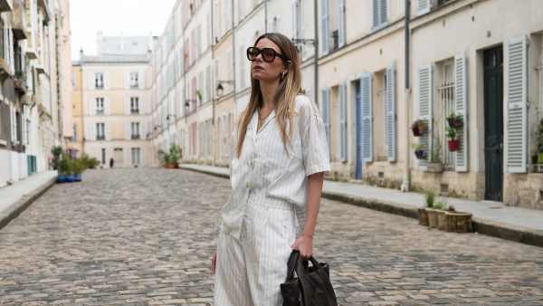 PARIS, FRANCE - MAY 07: Natalia Verza wears long straight light brown hair with blonde highlights and a center part, oversized black sunglasses with brown tinted lenses and thick frames, natural makeup with nude lipstick, a delicate silver-tone necklace, a white Munthe short-sleeve shirt with thin vertical gray stripes, an open collar, front button fastening, gold-tone buttons, a loose fit and a slightly uneven hem, white Munthe bermuda shorts with matching thin vertical gray stripes, a high waist, and knee-length cut, a dark brown leather Munthe bag with two short handles, a soft slouchy shape and tonal front logo detail, and brown Potamy slingback pumps with pointed toes, slim ankle straps, small kitten heels and a suede-effect finish, during a street style fashion photo session, on May 07, 2026 in Paris, France. (Photo by Edward Berthelot/Getty Images)