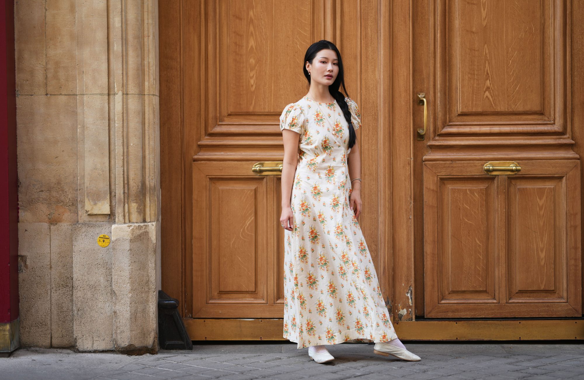PARIS, FRANCE - JUNE 14: Yuqing wears long black hair with a center part styled in a side braid, a cream maxi dress with short puff sleeves and an orange and green floral print, white ankle socks, a gold-tone bracelet, a metallic gold quilted leather shoulder bag, white pointed-toe ballet flats shoes, during a street style fashion photo session, on June 14, 2026 in Paris, France. (Photo by Edward Berthelot/Getty Images)