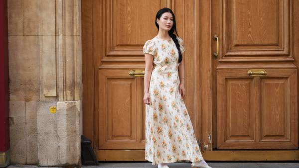 PARIS, FRANCE - JUNE 14: Yuqing wears long black hair with a center part styled in a side braid, a cream maxi dress with short puff sleeves and an orange and green floral print, white ankle socks, a gold-tone bracelet, a metallic gold quilted leather shoulder bag, white pointed-toe ballet flats shoes, during a street style fashion photo session, on June 14, 2026 in Paris, France. (Photo by Edward Berthelot/Getty Images)