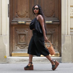 PARIS, FRANCE - JULY 19: Carrole Sagba wears long wavy black hair, black oval sunglasses, a black gathered cropped halterneck top by NA-KD, black culottes long shorts by Uniqlo, a natural wicker bucket bag by Parfois, black leather platform wedge sandals with wooden soles and gold-tone stud embellishments by Zara, gold-tone rings, during a street style fashion photo session, on July 19, 2026 in Paris, France. (Photo by Edward Berthelot/Getty Images)