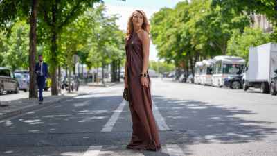 Chloé Lecareux wears a halter-neck dress in a rich brown satin fabric, featuring a flowing silhouette. The dress is complemented by a gold metallic collar detail. Hair is styled in loose waves. Accessories include a chunky bracelet with large, dark stones set in gold frames. A textured sequined brown handbag is carried, outside Stephane Rolland, during Paris Fashion Week - Haute Couture Fall/Winter 2025/2026, on July 08, 2025 in Paris, France