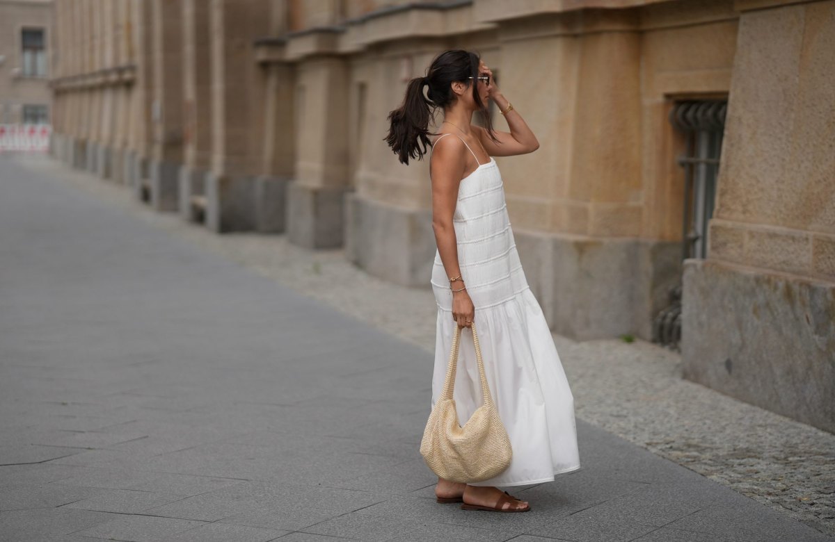 Aylin Sengül is seen wearing a white spaghetti-strap maxi dress with textured horizontal panels and a gathered hem by COS; brown leather “Oran” sandals by Hermès; a raffia shoulder bag with a slouchy hobo shape by COS; a delicate gold-tone necklace; a rose gold “Knot” bracelet by Celine; a vintage tennis bracelet; a slim silver-tone bangle; a gold ring; slim oval dark tortoiseshell sunglasses by Chanel during Berlin Fashion Week Spring/Summer 2027 on July 4, 2026 in Berlin, Germany.