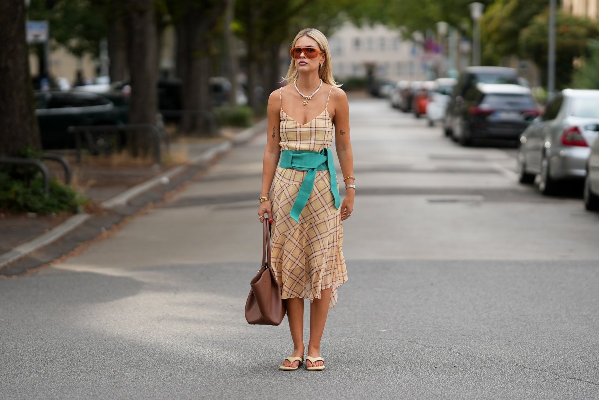 Julia Kammerer is seen wearing a beige plaid midi dress from Mango; a wide turquoise fabric belt from Mango; beige flip flops from Ulanka; a large brown leather tote bag with gold tone hardware from DeMellier; layered pearl and gold chain necklaces from Chanel and Akind & Scandiv, gold hoop earrings, rings from Pandora, Scandiv and Akind, gold bracelets and a silver and gold tone watch from Mockberg; oversized translucent acetate sunglasses with brown tinted lenses from & Other Stories; blonde hair worn straight with a center part on August 7, 2026 in Frankfurt am Main, Germany.