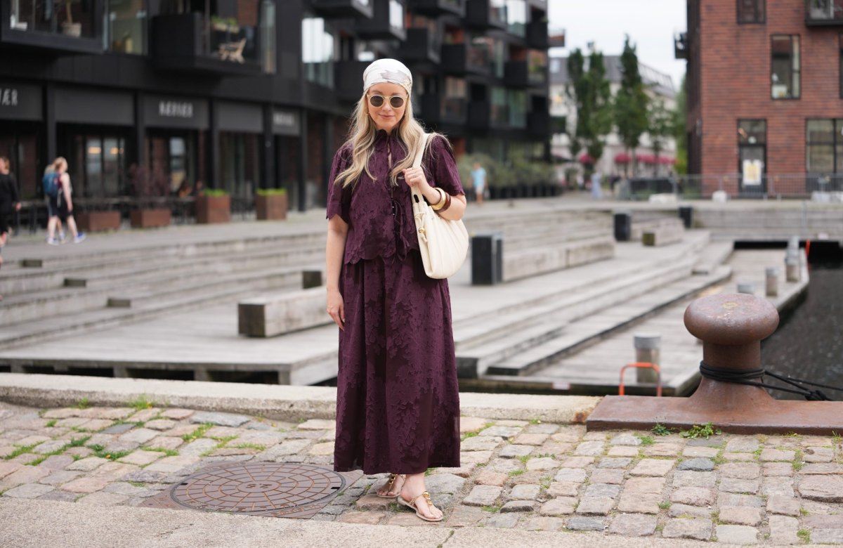 COPENHAGEN, DENMARK - AUGUST 04: A guest wears a white and beige printed headscarf, long wavy blonde hair, round sunglasses with dark lenses, a dark purple short-sleeve top with floral pattern and tie-front detailing, a matching maxi skirt, forming a maxi dress shape, a beige leather shoulder bag, sandals shoes, bangles, outside Rene, during Copenhagen Fashion Week Spring/Summer 2027, on August 04, 2026 in Copenhagen, Denmark (Photo by Edward Berthelot/Getty Images)