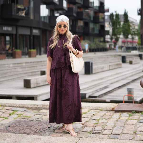 COPENHAGEN, DENMARK - AUGUST 04: A guest wears a white and beige printed headscarf, long wavy blonde hair, round sunglasses with dark lenses, a dark purple short-sleeve top with floral pattern and tie-front detailing, a matching maxi skirt, forming a maxi dress shape, a beige leather shoulder bag, sandals shoes, bangles, outside Rene, during Copenhagen Fashion Week Spring/Summer 2027, on August 04, 2026 in Copenhagen, Denmark (Photo by Edward Berthelot/Getty Images)