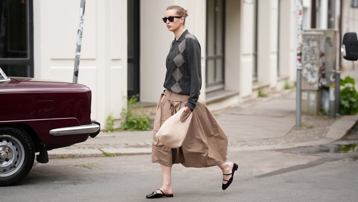 BERLIN, GERMANY - MAY 15: Sina Anjulie is seen wearing a dark charcoal grey argyle knit jacket with a spread polo collar, button-front closure and long sleeves from COS; a camel colored "Pleated Palloncino" midi skirt with deep box pleats, a wide structured waistband and a voluminous balloon silhouette in a lightweight cotton from Litkovska; black patent leather "Jane Nina" flat mules with a double Mary Jane strap detail from Christian Louboutin; a cream knitted bag with a rounded silhouette and short looped top handles from &Other Stories; a delicate hoop earring stack; "Wayfarer" sunglasses with a black frame and dark tinted lenses from Ray-Ban; her brown hair is slicked back into a low bun; with a natural make-up on May 15, 2026 in Berlin, Germany. (Photo by Moritz Scholz/Getty Images)