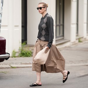BERLIN, GERMANY - MAY 15: Sina Anjulie is seen wearing a dark charcoal grey argyle knit jacket with a spread polo collar, button-front closure and long sleeves from COS; a camel colored "Pleated Palloncino" midi skirt with deep box pleats, a wide structured waistband and a voluminous balloon silhouette in a lightweight cotton from Litkovska; black patent leather "Jane Nina" flat mules with a double Mary Jane strap detail from Christian Louboutin; a cream knitted bag with a rounded silhouette and short looped top handles from &Other Stories; a delicate hoop earring stack; "Wayfarer" sunglasses with a black frame and dark tinted lenses from Ray-Ban; her brown hair is slicked back into a low bun; with a natural make-up on May 15, 2026 in Berlin, Germany. (Photo by Moritz Scholz/Getty Images)
