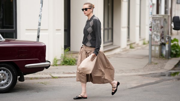 BERLIN, GERMANY - MAY 15: Sina Anjulie is seen wearing a dark charcoal grey argyle knit jacket with a spread polo collar, button-front closure and long sleeves from COS; a camel colored "Pleated Palloncino" midi skirt with deep box pleats, a wide structured waistband and a voluminous balloon silhouette in a lightweight cotton from Litkovska; black patent leather "Jane Nina" flat mules with a double Mary Jane strap detail from Christian Louboutin; a cream knitted bag with a rounded silhouette and short looped top handles from &Other Stories; a delicate hoop earring stack; "Wayfarer" sunglasses with a black frame and dark tinted lenses from Ray-Ban; her brown hair is slicked back into a low bun; with a natural make-up on May 15, 2026 in Berlin, Germany. (Photo by Moritz Scholz/Getty Images)