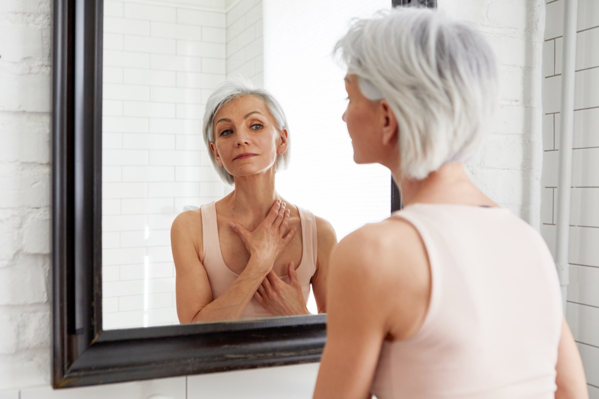 Beautiful senior woman morning bathroom routine, looks into mirror touches her neck with sensual movement of hands. Elderly female natural beauty. Dignity and grace in old age. Over the Shoulder Shot