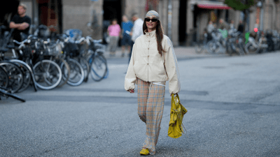 Evi Wave wears brown Prada sunglasses, a yellow and blue checked Reclaimed Vintage headscarf, a cream Topshop bomber jacket with stand collar and frog closures, sheer yellow and blue checked Reclaimed Vintage trousers, yellow and black Onitsuka Tiger sneakers and carries a yellow Balenciaga City bag with mirror detail outside the Caro Editions show during day 4 of Copenhagen Fashion Week Spring Summer 2027 on August 6, 2026 in Copenhagen, Denmark.