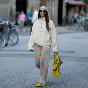 Evi Wave wears brown Prada sunglasses, a yellow and blue checked Reclaimed Vintage headscarf, a cream Topshop bomber jacket with stand collar and frog closures, sheer yellow and blue checked Reclaimed Vintage trousers, yellow and black Onitsuka Tiger sneakers and carries a yellow Balenciaga City bag with mirror detail outside the Caro Editions show during day 4 of Copenhagen Fashion Week Spring Summer 2027 on August 6, 2026 in Copenhagen, Denmark.