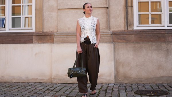 COPENHAGEN, DENMARK - AUGUST 05: A guest wears slicked back dark brown hair, a white lace sleeveless mandarin collar top with frog closures and floral embroidery, brown pleated wide-leg balloon pants, a black leather belt, a dark green leather top-handle bag, black open-toe kitten heel sandals shoes, outside The Garment, during Copenhagen Fashion Week Spring/Summer 2027, on August 05, 2026 in Copenhagen, Denmark (Photo by Edward Berthelot/Getty Images)