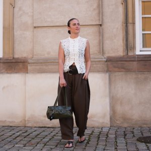 COPENHAGEN, DENMARK - AUGUST 05: A guest wears slicked back dark brown hair, a white lace sleeveless mandarin collar top with frog closures and floral embroidery, brown pleated wide-leg balloon pants, a black leather belt, a dark green leather top-handle bag, black open-toe kitten heel sandals shoes, outside The Garment, during Copenhagen Fashion Week Spring/Summer 2027, on August 05, 2026 in Copenhagen, Denmark (Photo by Edward Berthelot/Getty Images)
