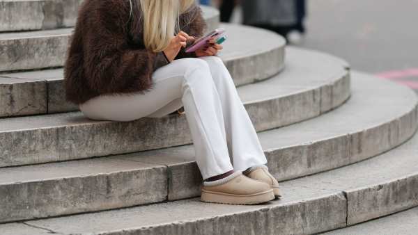 A passerby wears blond long hair, a brown fluffy faux fur oversized winter jacket / coat, white pants, beige UGG suede shoes with thick rubber soles, uses a pink mobile phone / smart phone, during a street style fashion photo session, on December 13, 2025 in Paris, France.