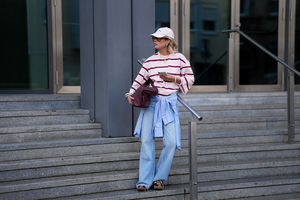 FRANKFURT AM MAIN, GERMANY - MARCH 26: Julia Kammerer is seen wearing an oversized pink and burgundy horizontal striped cotton T shirt from H&M; a blue and white striped button down shirt tied around her waist from JJXX; light blue wide leg denim jeans with a relaxed fit from JJXX; burgundy and yellow textured leather lace up sneakers from Adidas; a large burgundy woven leather hobo bag from NA-KD; a pale pink baseball cap with the red embroidered text "Amore a Milano" from LeGer by Lena Gercke; her jewelry includes chunky gold hoop earrings, necklaces and gold tone stacked rings worn across multiple fingers from H&M, Pandora and My Jewellery; holding an iPhone from Apple; several silver and resin bangles from & Other Stories; orange tinted aviator sunglasses with a thin black frame from Bershka; her blonde hair is styled in loose waves underneath the cap on March 26, 2026 in Frankfurt am Main, Germany. (Photo by Moritz Scholz/Getty Images)