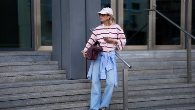 FRANKFURT AM MAIN, GERMANY - MARCH 26: Julia Kammerer is seen wearing an oversized pink and burgundy horizontal striped cotton T shirt from H&M; a blue and white striped button down shirt tied around her waist from JJXX; light blue wide leg denim jeans with a relaxed fit from JJXX; burgundy and yellow textured leather lace up sneakers from Adidas; a large burgundy woven leather hobo bag from NA-KD; a pale pink baseball cap with the red embroidered text "Amore a Milano" from LeGer by Lena Gercke; her jewelry includes chunky gold hoop earrings, necklaces and gold tone stacked rings worn across multiple fingers from H&M, Pandora and My Jewellery; holding an iPhone from Apple; several silver and resin bangles from & Other Stories; orange tinted aviator sunglasses with a thin black frame from Bershka; her blonde hair is styled in loose waves underneath the cap on March 26, 2026 in Frankfurt am Main, Germany. (Photo by Moritz Scholz/Getty Images)