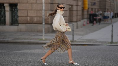 Lisa Mottl is seen wearing an off white high neck oversized windbreaker jacket with elasticated cuffs from Arket; a brown, grey and mustard plaid asymmetrical flowing midi skirt from Zara; silver metallic pointed toe slingback kitten heels from H&M; a beige crocodile embossed structured clutch bag from H&M; black rectangular frame sunglasses from Vehla; silver hoop earrings, silver rings on July 23, 2026 in Berlin, Germany.