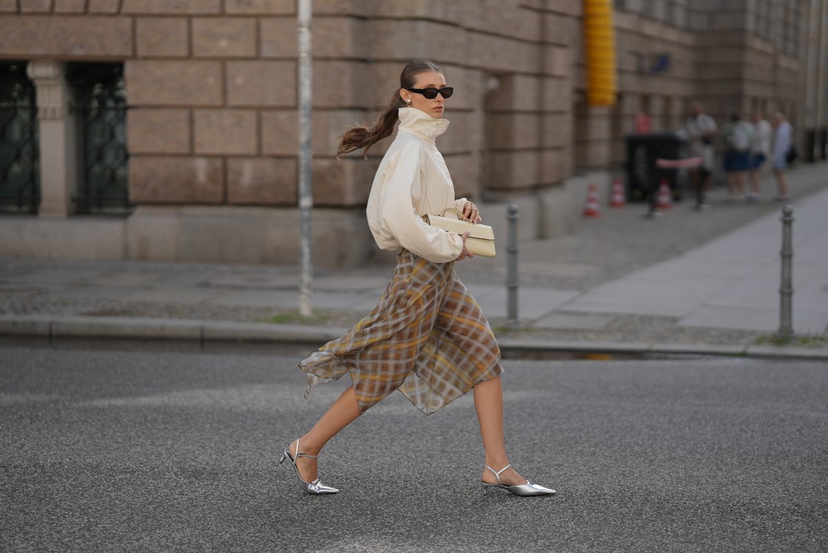 Lisa Mottl is seen wearing an off white high neck oversized windbreaker jacket with elasticated cuffs from Arket; a brown, grey and mustard plaid asymmetrical flowing midi skirt from Zara; silver metallic pointed toe slingback kitten heels from H&M; a beige crocodile embossed structured clutch bag from H&M; black rectangular frame sunglasses from Vehla; silver hoop earrings, silver rings on July 23, 2026 in Berlin, Germany.