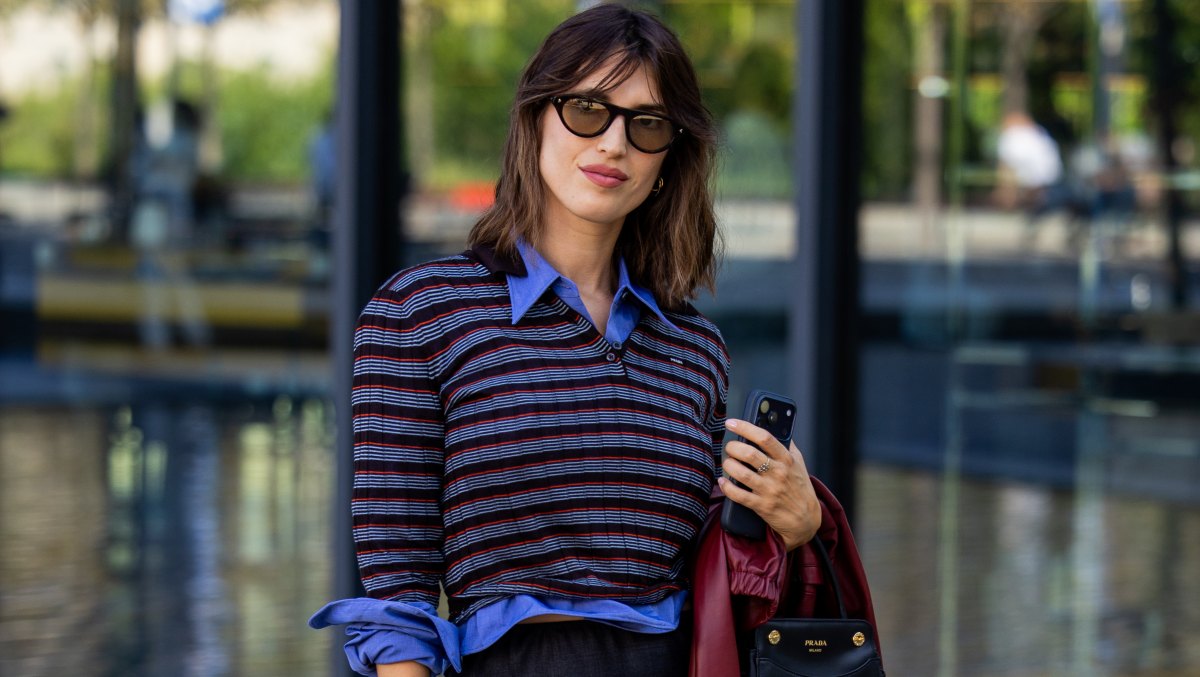 MILAN, ITALY - SEPTEMBER 22: Jeanne Damas wears a striped long-sleeved polo shirt, a blue collared shirt, a grey pencil skirt, black pointed-toe slingback heels, sunglasses, a red jacket and a black "PRADA" handbag outside Prada on September 22, 2026 in Milan, Italy. (Photo by Christian Vierig/Getty Images)