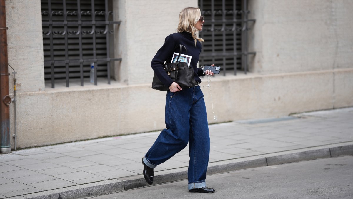 MUNICH, GERMANY - FEBRUARY 18: Mirja Klein is seen wearing a navy fine knit turtleneck sweater with long sleeves and a fitted silhouette from COS; dark wash baggy jeans with a relaxed oversized barrel fit and rolled-up legs from Dark Park; a slim black leather belt with silver buckle; black polished leather loafers with a sleek rounded toe and slim profile from COS; a black „Aventure“ nappa leather bag with gold hardware, chain link details and a belted detail in the front closure from Miu Miu; oversized shield sunglasses with a thick transparent acetate frame and brown tinted lenses from Bottega Veneta; oversized brown shield sunglasses with dark tinted lenses and a thick glossy frame from MM6 Maison Margiela; holding an iPhone smartphone and wearing white wired earphones; her blonde hair is worn long and wavy with a center part on February 18, 2026 in Munich, Germany. (Photo by Moritz Scholz/Getty Images)