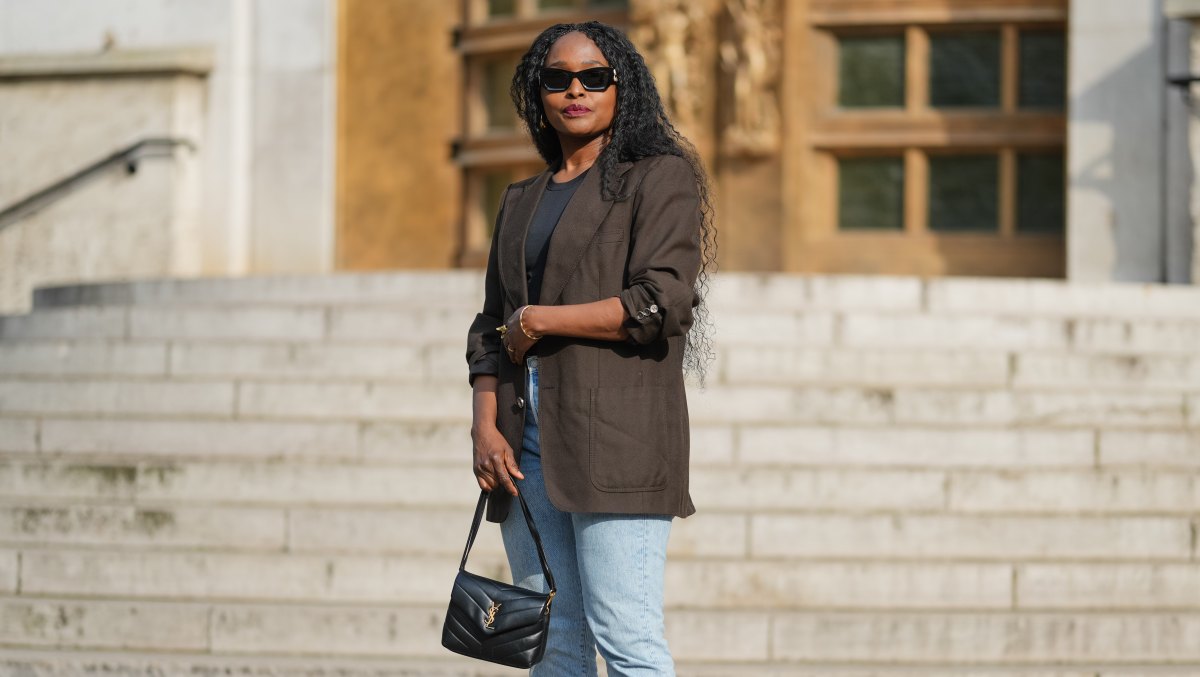 PARIS, FRANCE - MAY 02: Carrole Sagba wears long, curly black hair parted in the middle. Black sunglasses cover the eyes, and red lipstick is applied. A dark brown blazer jacket made of a textured material is worn over a black top. Light blue denim jeans with a straight cut are paired with black studded slip-on shoes. A black leather YSL Saint Laurent handbag with a visible gold logo is carried. The image is a full body shot, during a street style fashion photo session, on May 02, 2025 in Paris, France. (Photo by Edward Berthelot/Getty Images)