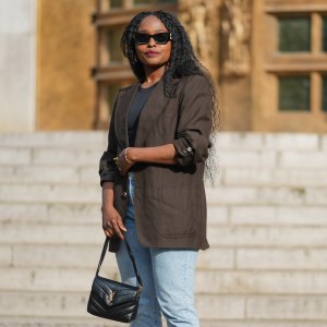 PARIS, FRANCE - MAY 02: Carrole Sagba wears long, curly black hair parted in the middle. Black sunglasses cover the eyes, and red lipstick is applied. A dark brown blazer jacket made of a textured material is worn over a black top. Light blue denim jeans with a straight cut are paired with black studded slip-on shoes. A black leather YSL Saint Laurent handbag with a visible gold logo is carried. The image is a full body shot, during a street style fashion photo session, on May 02, 2025 in Paris, France. (Photo by Edward Berthelot/Getty Images)