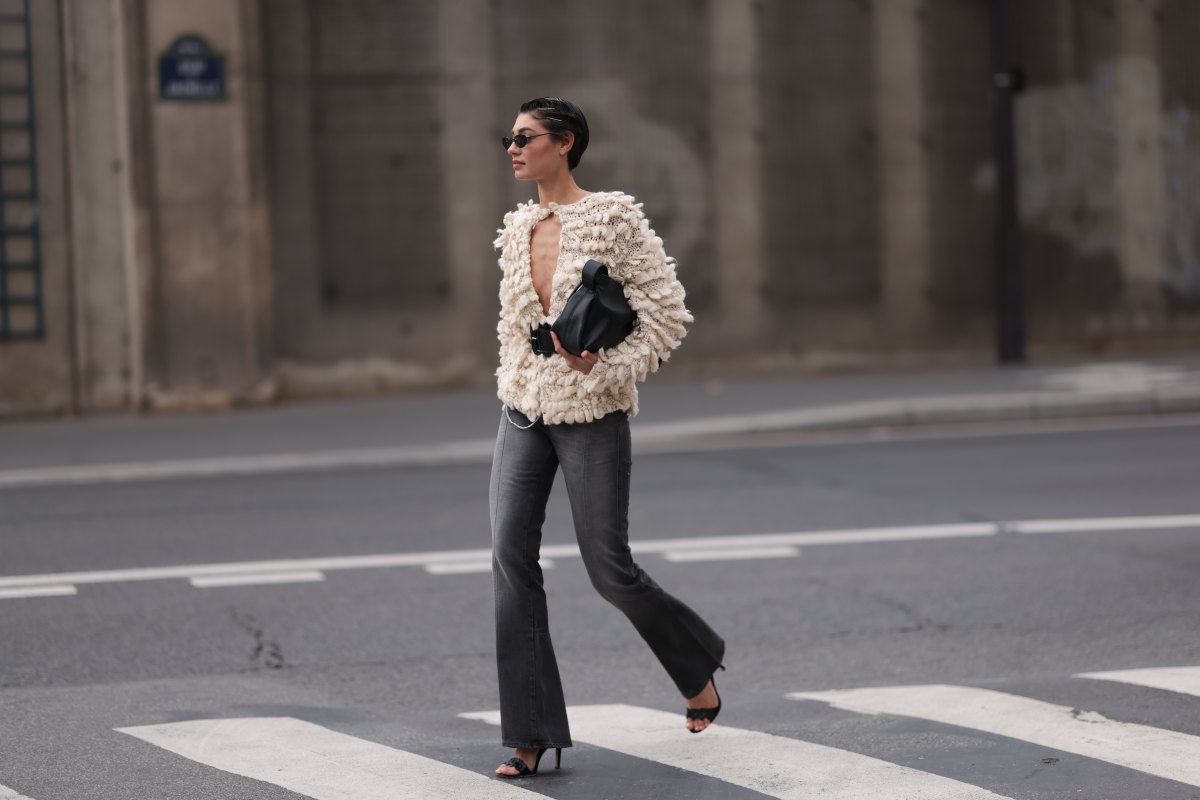 Sarah Posch is seen wearing rimless oval sunglasses with thin black metal temples and nose bridge, a beige chunky-knitted cardigan with fluffy rounded short fringes, a wide black leather belt with oversized black buckles, a black leather bag, slightly faded grey jeans with decorative seams, and black leather heeled sandals before the Ottolinger Womenswear Spring/Summer 2025 Show as part of Paris Fashion Week on September 29, 2024 in Paris, France.