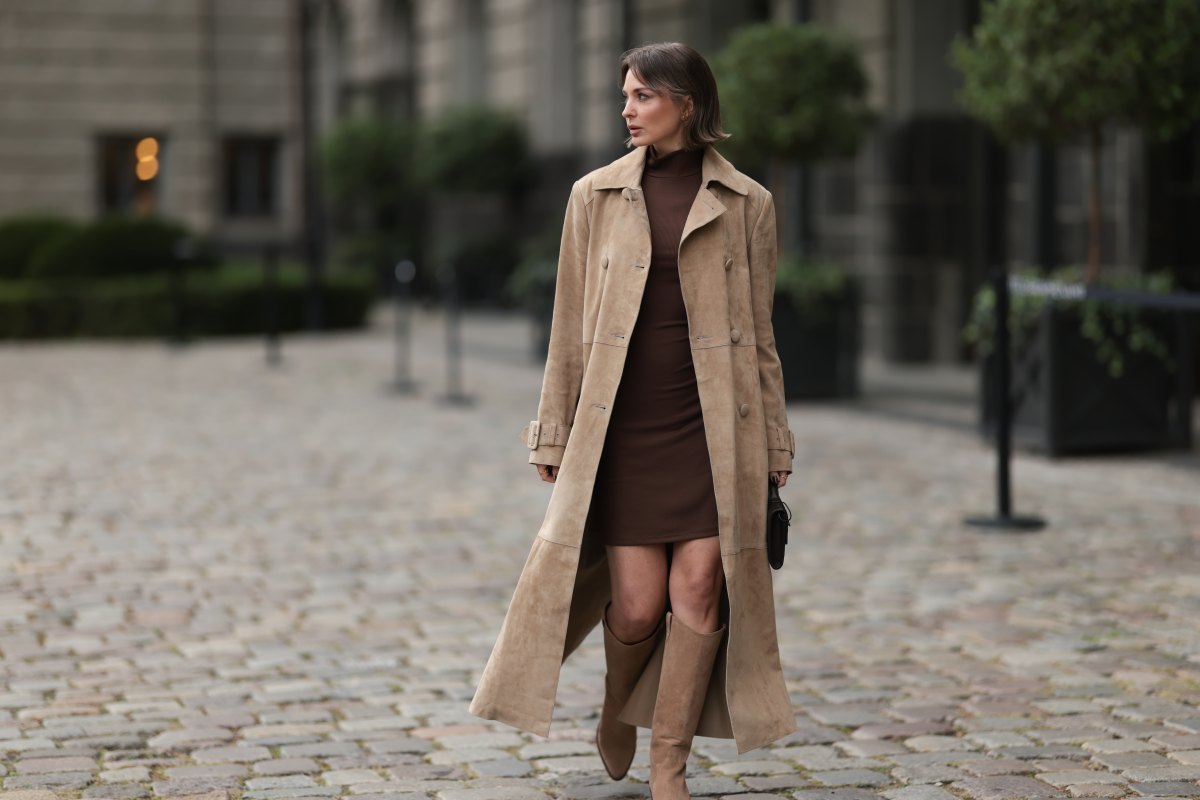 Yana Pries seen wearing a long suede leather trench coat from NOUR HAMMOUR in a warm beige tone. The coat features double-breasted buttons, a structured collar, and belted cuffs, creating a luxurious yet timeless silhouette. Her hair is cut into a sleek bob, light brown with subtle highlights, styled with a middle part and softly curved ends that frame her face. Underneath she wears Even&Odd fitted chocolate-brown mini dress, with a high neckline, Bottega Veneta Andiamo clutch, a structured black leather bag with a gold knot detail on the strap and Bobbies knee-high Bobbies suede leather boots in a soft beige tone, on September 17, 2025 in Berlin, Germany.