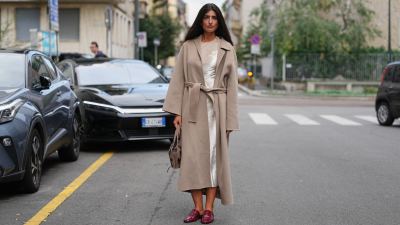 A guest wears long dark brown hair worn loose with a center part, silver stud earrings, a taupe suede hobo bag with dark floral embroidery, a calf-length belted camel-beige wool coat with a wide notched collar and long sleeves, layered over an ivory satin slip dress with visible sheen at the neckline and hem, pointed-toe burgundy patent leather backless mules with a low stacked heel, outside Alberta Ferretti, during Milan Fashion Week - WoWomenswear Spring/Summer 2026, on September 23, 2025 in Milan, Italy