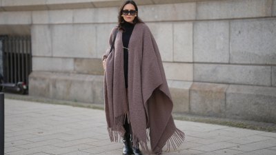 Laura Noltemeyer is seen wearing an oversized fringed wool poncho in a mauve tone with a textured, fluffy finish, fringes at the hem and a gold pin closure element called „Poncho COZY“ by Aylin Koenig; black fine-knit turtleneck sweater with slim-fitting silhouette by Aylin Koenig; knee-high black leather boots with a rounded toe and flat sole by Massimo Dutti; a structured black leather bucket bag with a long shoulder strap and gold hardware details by Sézane; among her jewelry is a white gold „LOVE“ bracelet by Cartier; oversized square black acetate sunglasses with ombré tinted lenses by Céline; large gold statement earrings with a ruffled surface and a green stone element by Mango; her dark brown hair is styled in loose waves with a center part on November 13, 2025 in Hamburg, Germany.