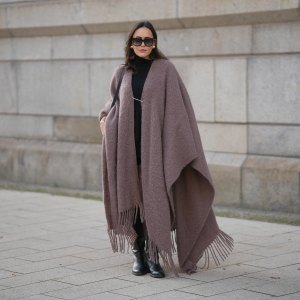 Laura Noltemeyer is seen wearing an oversized fringed wool poncho in a mauve tone with a textured, fluffy finish, fringes at the hem and a gold pin closure element called „Poncho COZY“ by Aylin Koenig; black fine-knit turtleneck sweater with slim-fitting silhouette by Aylin Koenig; knee-high black leather boots with a rounded toe and flat sole by Massimo Dutti; a structured black leather bucket bag with a long shoulder strap and gold hardware details by Sézane; among her jewelry is a white gold „LOVE“ bracelet by Cartier; oversized square black acetate sunglasses with ombré tinted lenses by Céline; large gold statement earrings with a ruffled surface and a green stone element by Mango; her dark brown hair is styled in loose waves with a center part on November 13, 2025 in Hamburg, Germany.