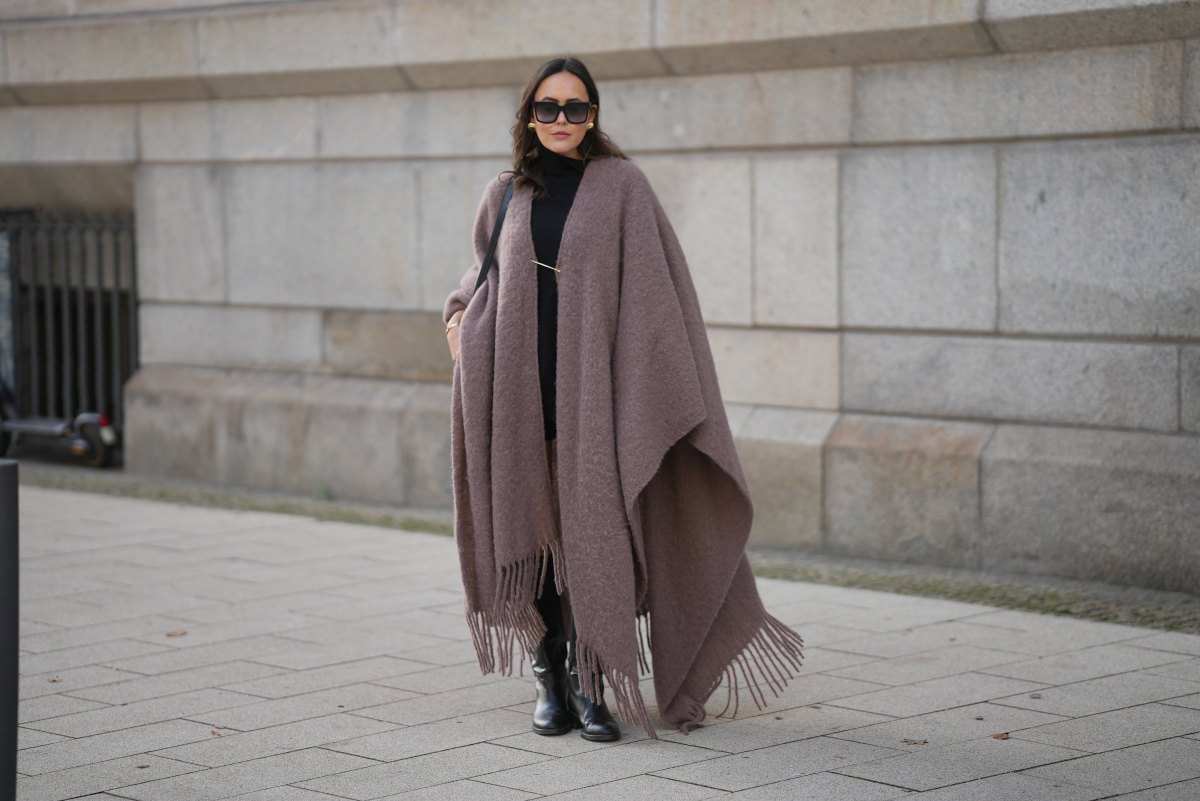 Laura Noltemeyer is seen wearing an oversized fringed wool poncho in a mauve tone with a textured, fluffy finish, fringes at the hem and a gold pin closure element called „Poncho COZY“ by Aylin Koenig; black fine-knit turtleneck sweater with slim-fitting silhouette by Aylin Koenig; knee-high black leather boots with a rounded toe and flat sole by Massimo Dutti; a structured black leather bucket bag with a long shoulder strap and gold hardware details by Sézane; among her jewelry is a white gold „LOVE“ bracelet by Cartier; oversized square black acetate sunglasses with ombré tinted lenses by Céline; large gold statement earrings with a ruffled surface and a green stone element by Mango; her dark brown hair is styled in loose waves with a center part on November 13, 2025 in Hamburg, Germany.