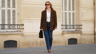 Merve Gorgoz wears brown long hair with center part, amber colored sunglasses with orange tinted lenses by Jimmy Fairly, golden square earrings, a brown suede oversize blazer jacket by Source Unknown, a white shirt, a leather belt, blue denim pants jeans by Mango, a dark brown khaki bag in suede by Demellier, brown pointed shiny patent leather shoes with high heels by YSL Saint Laurent, during a street style fashion photo session, on September 01, 2026 in Paris, France.