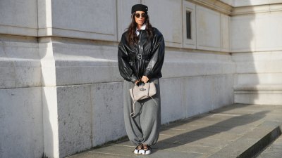 PARIS, FRANCE - DECEMBER 09: Gabriella Berdugo wears long dark brown hair worn loose under a black pillbox hat with stud details by Gabriella Berdugo Studio, natural skin with neutral lipstick, dark tinted sunglasses, a white button front shirt by Mum Of and a tie, a black leather bomber style jacket with dropped shoulders, elasticated cuffs, zip front closure and a matching leather belt by The Frankie Shop, a black tie by Prada worn at the neckline, grey sarouel style trousers by Zara with a loose tapered silhouette, white and black ballet flat shoes by Adidas with contrast striping, and a beige leather top handle bag by Longchamp, during a street style fashion photo session, on December 09, 2025 in Paris, France. (Photo by Edward Berthelot/Getty Images)