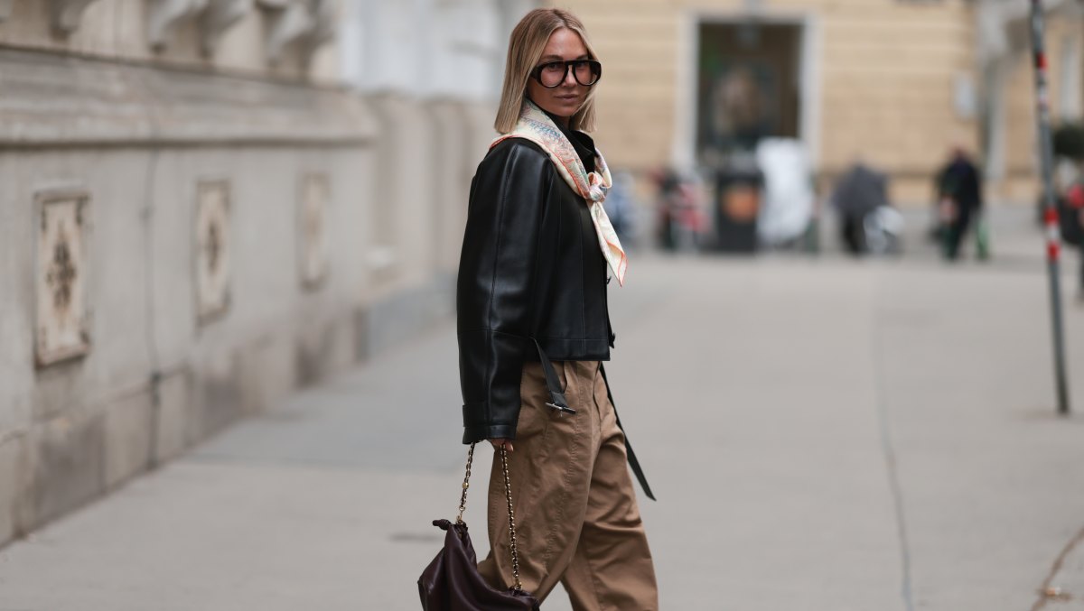 VIENNA, AUSTRIA - MARCH 16: Karin Teigl is seen wearing a Zara orange crew-neck t-shirt, a Loewe black leather jacket with a relaxed fit, loose-fitting high-waisted Uniqlo brown trousers with prominent seams and pleats, and Miu Miu x New Balance light-colored sneakers with white laces. She also wears Andy Wolf oversized black glasses, a patterned Hermes silk scarf tied around her neck, a delicate green pendant necklace, and carries a small dark brown gathered Loewe clutch bag with a gold chain strap, with Miu Miu underwear slightly visible above her trousers on March 16, 2026 in Vienna, Austria. (Photo by Jeremy Moeller/Getty Images)