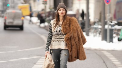 sweater vest Evi Wave is seen wearing a voluminous brown faux fur jacket with an oversized silhouette and wide lapels from Munthe; a cream sequin embellished knit vest with black geometric pattern detailing from Munthe; a black and white striped long sleeve top in a fitted silhouette from Munthe; grey wide leg jeans adorned with multicolored floral crystal embellishments and a bootcut shape from Munthe; black leather pointed toe ankle boots featuring a slim heel from Munthe; a beige padded clutch bag crafted in softly ruched textured fabric with an imprinted pattern, drawstring details and a keyring charm with brand tag from Munthe; a brown knitted beanie from Munthe; silver ring stack jewelry; her brown straight hair worn loose and straight on January 6, 2026 in Hamburg, Germany. (Photo by Moritz Scholz/Getty Images)